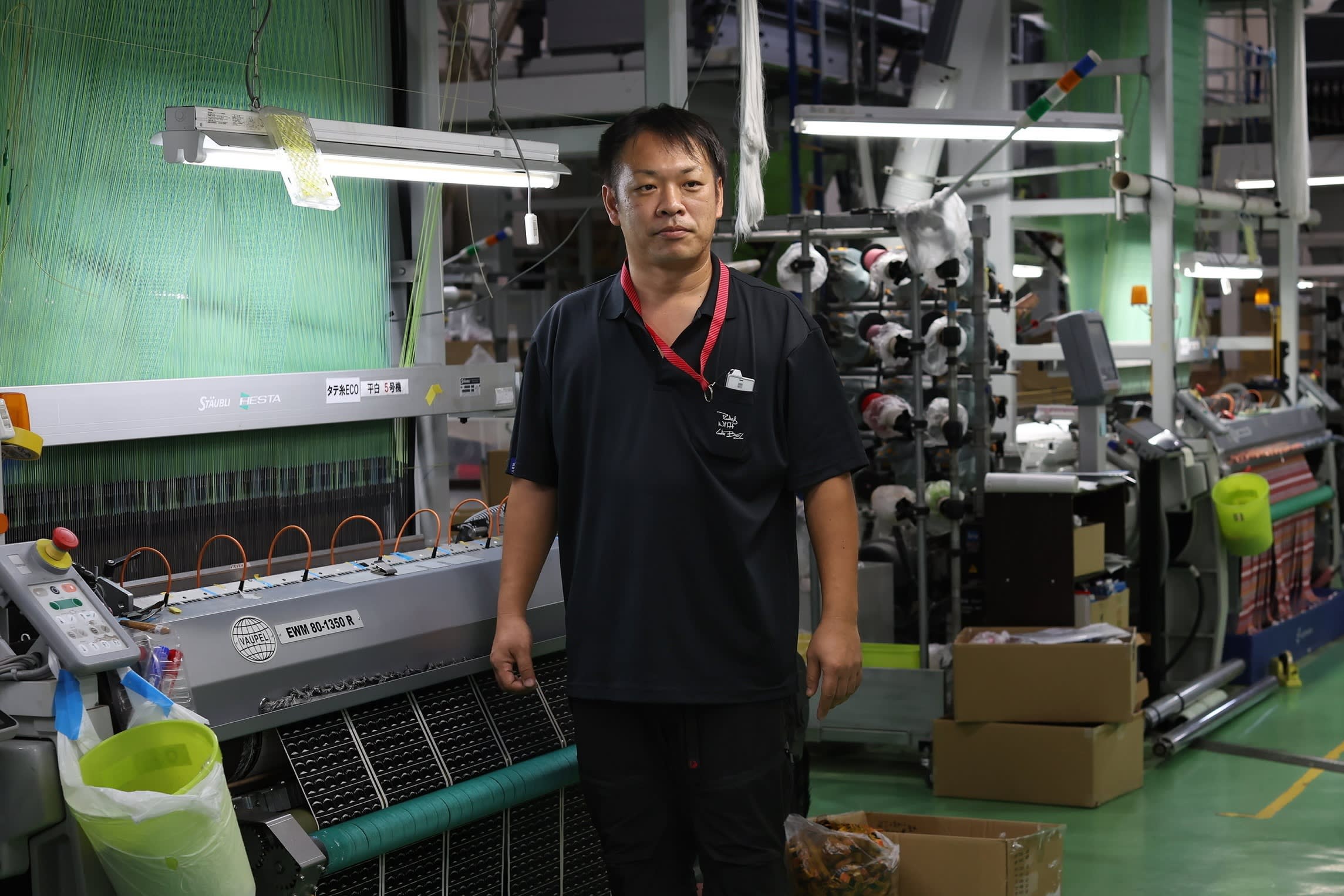Makoto Monzen stands on a factory floor at Matsukawa Rapyarn, surrounded by textile machinery and weaving equipment.