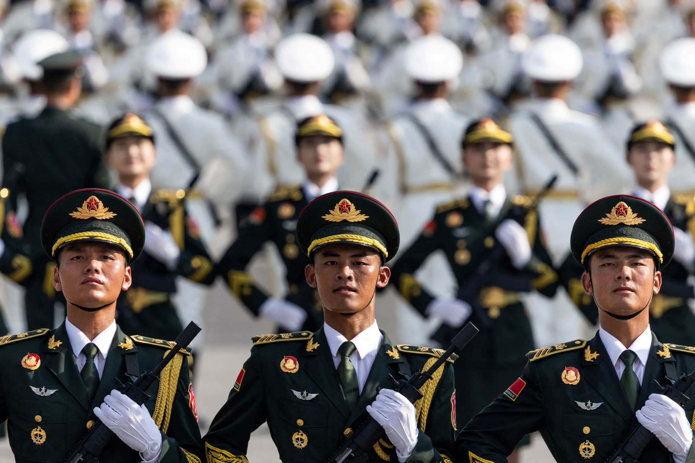 Three uniformed Chinese PLA soldiers stand at attention in the foreground with rows of soldiers behind them during a training exercise.