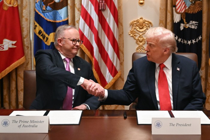 Anthony Albanese and Donald Trump shake hands across a table after signing an agreement, with flags in the background.