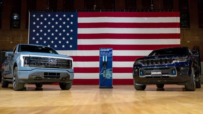 Ford F-150 Lightning and Jeep Grand Cherokee 4xe electric vehicles displayed with a charging station, large US flag in background.