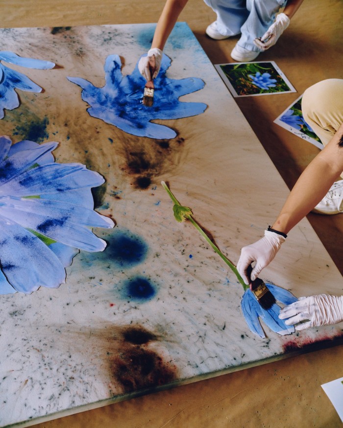 Photograph of two people painting blue flowers on paper stuck to a stretched canvas
