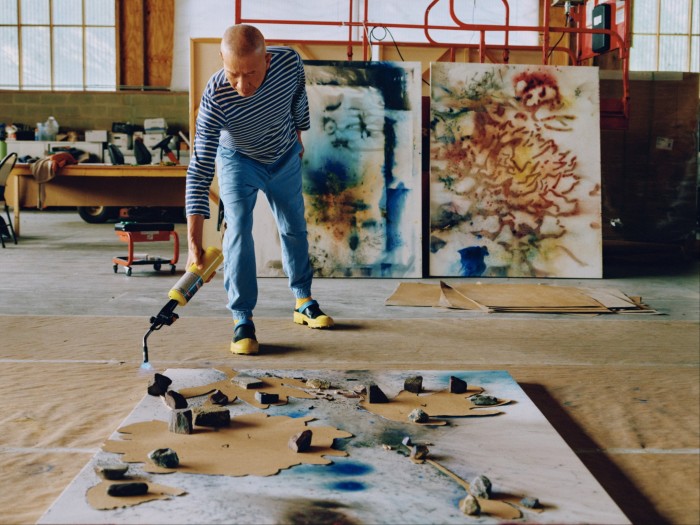 A stretched canvas is laid out on the wooden floor of an artist’s studio, laden with what looks like rocks and sprinklings of gunpowder, while a man sets light to the objects with a blowtorch
