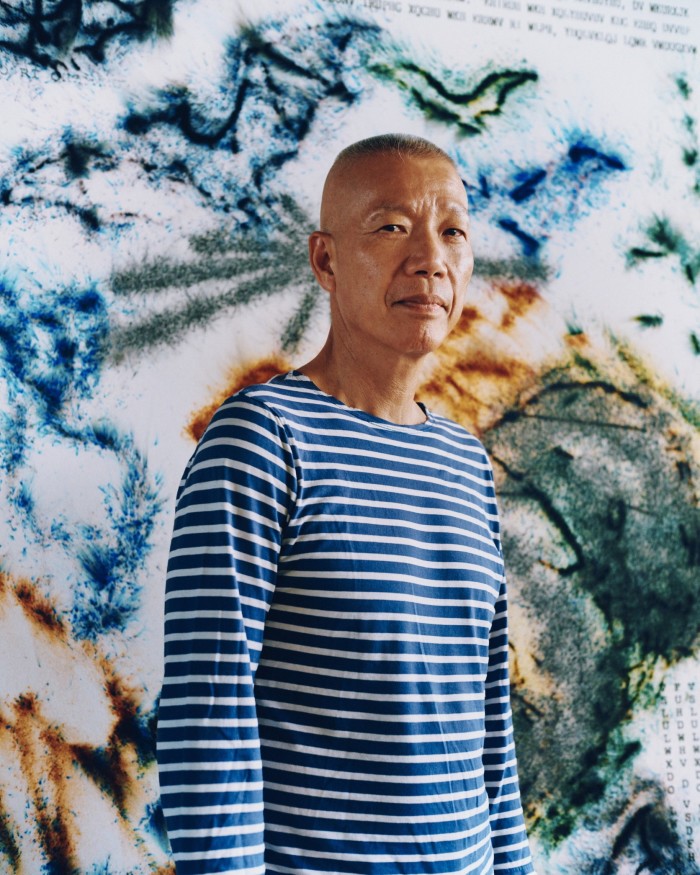 A middle-aged man with a shaved head, wearing a blue-and-white striped Breton top, stands in an artist’s studio