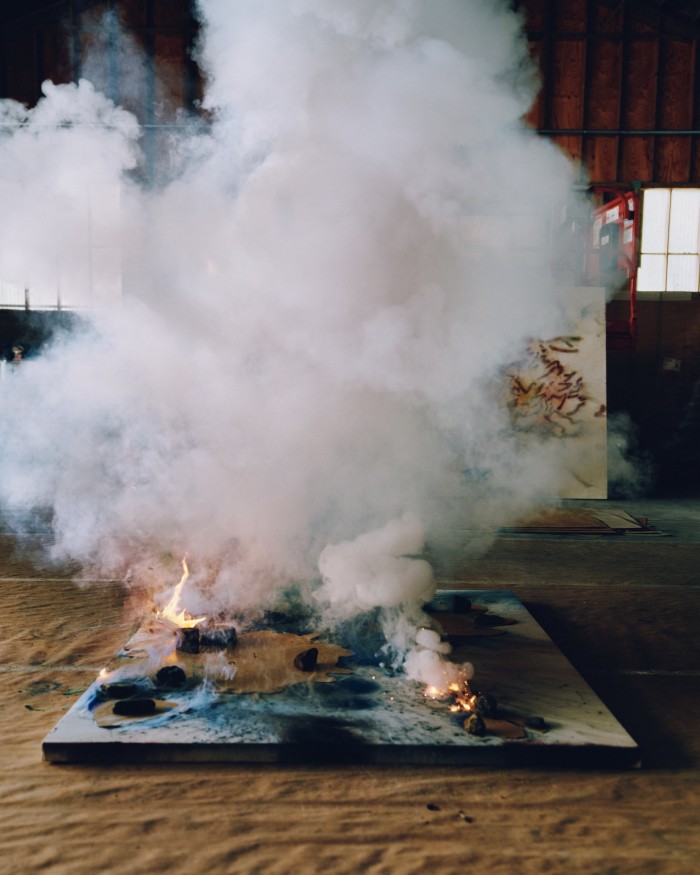 A stretched canvas laden with paint and explosives lies on the wooden floor of an artist’s studio, with flames and smoke rising from its surface. 