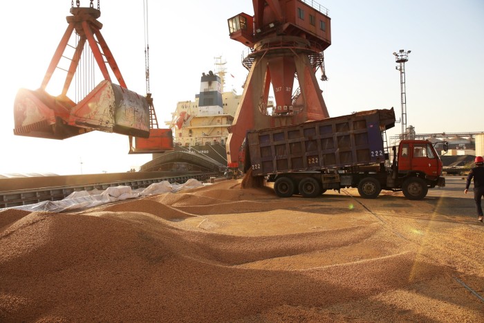 Soybeans being unloaded from a truck onto the ground at a port, with a crane and cargo ship in the background.