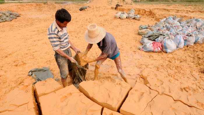 Two labourers fill a sack with yellow earth at a rare earth metals mine, with piles of filled sacks in the background.
