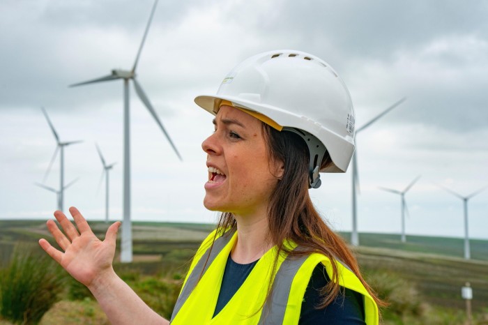 Kate Forbes wearing a hard hat and high-visibility vest, gesturing while speaking in front of several wind turbines at a wind farm.