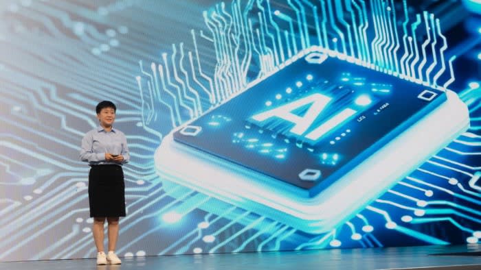 Yi Ying stands on stage speaking in front of a large digital display featuring an AI microchip graphic with glowing circuit lines.