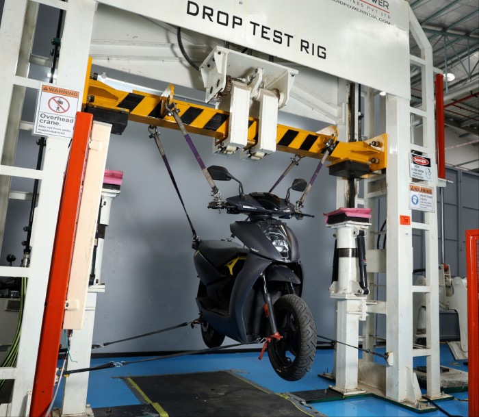 A black Ather electric scooter is suspended by straps in a drop test rig