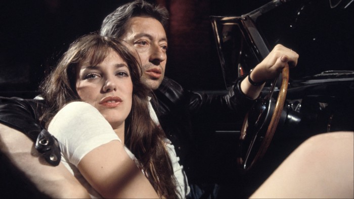 Jane Birkin and Serge Gainsbourg sit closely together in a car, Birkin leaning against Gainsbourg, who is holding the steering wheel.