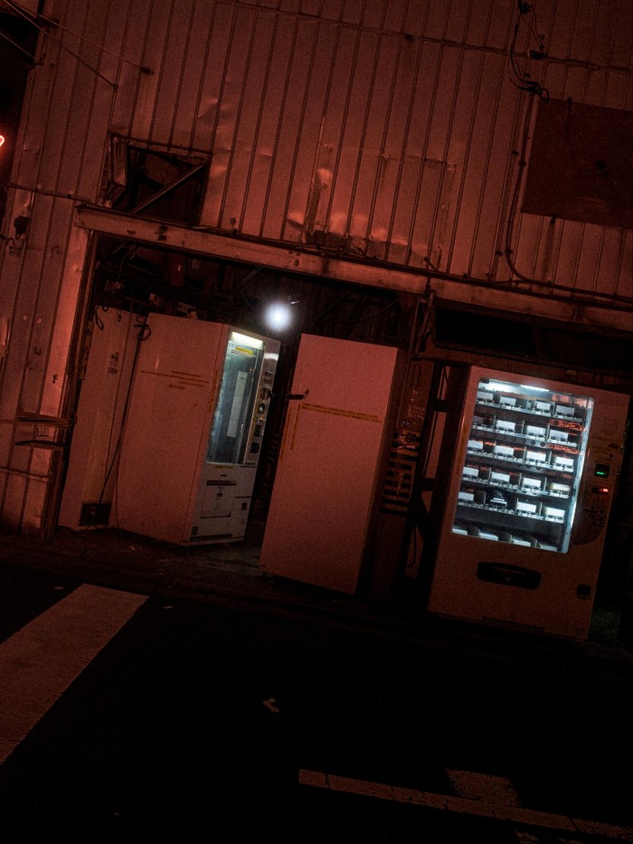 Two vending machines stand outside a dimly lit, industrial-looking building in Akhibara at night, illuminated by eerie red and white lights.