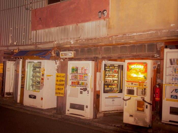 Cut-outs of two disembodied heads on the wall above a row of the old vending machines at night.