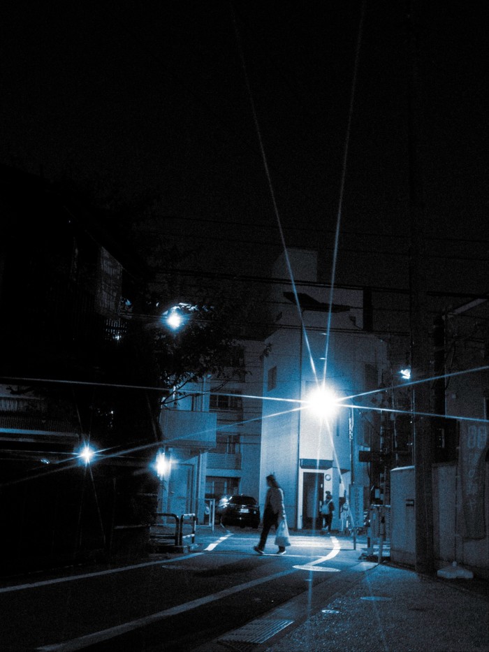 At night, a person carrying a bag walks down a dimly lit street by the Myogyo-ji Temple which is named after Oiwa, the ghost whose grave is found in its cemetery. The street is illuminated by street lights near residential buildings.
