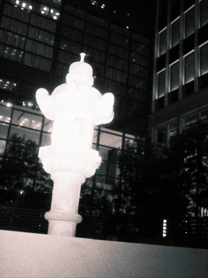 The grave of Taira no Masakado brightly at night, set against the glass facades of tall modern skyscrapers.