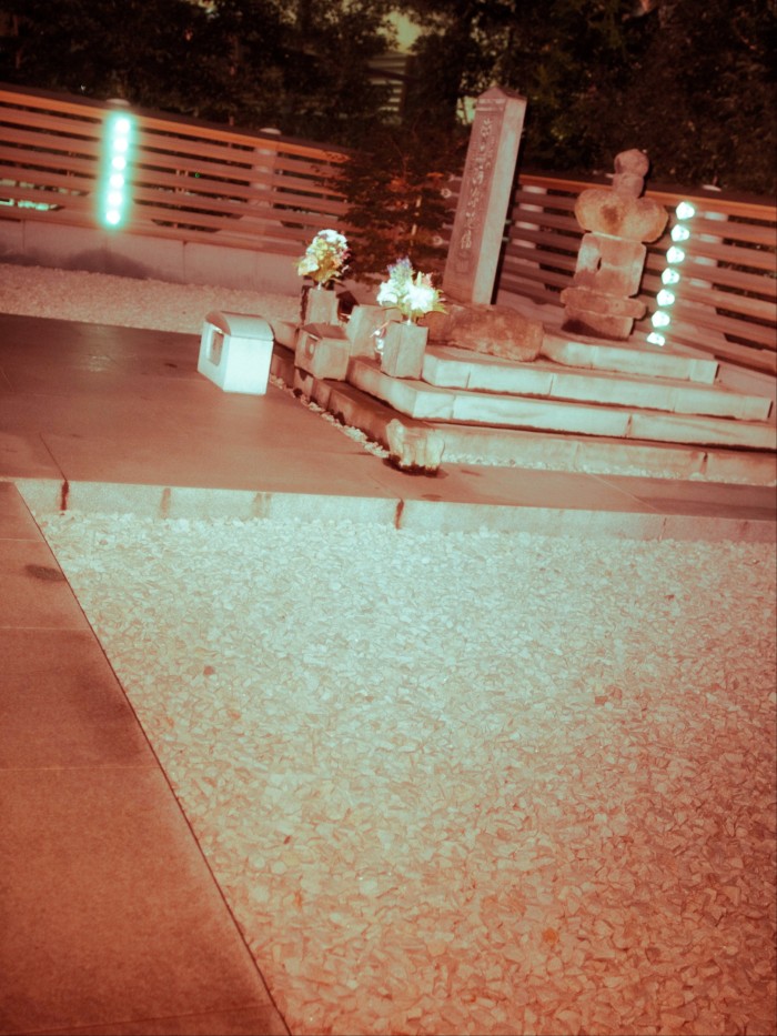 Stone grave marker for Taira no Masakado, with flowers placed at the base and illuminated by lights at night.