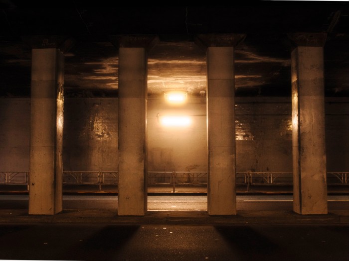 Five concrete pillars in Tokyo’s Sendagaya Tunnel, illuminated by a single central light casting warm yellow tones and deep shadows.