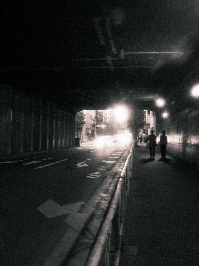 Two people walk on the sidewalk through the dimly lit Sendagaya tunnel in Tokyo, with car headlights shining towards the exit.