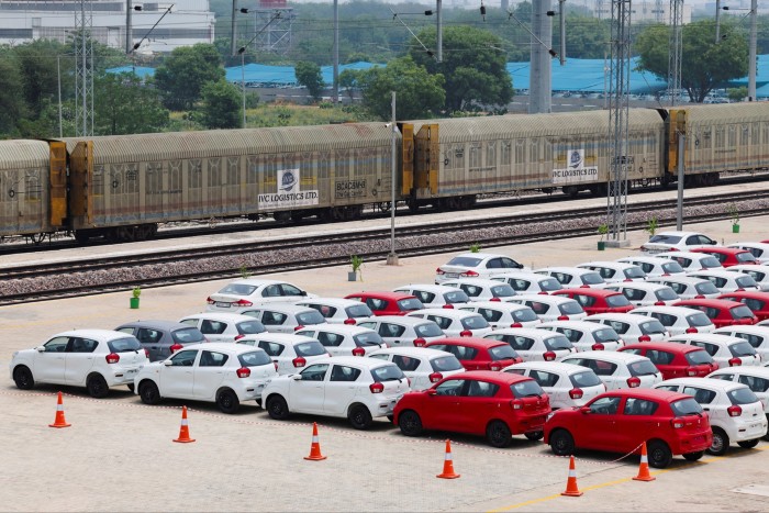 Rows of newly manufactured Maruti Suzuki Celerio cars, mostly white with some red, parked by a railway siding near a freight train.