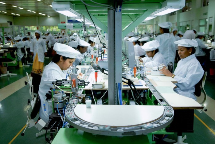 Rows of workers in a brightly lit factory, wearing white coats and caps, focus on assembling small components along a mechanized production line.