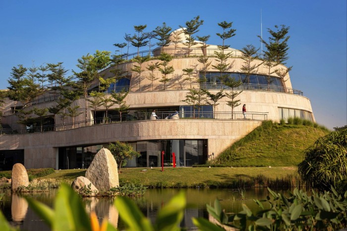 A modern circular building with trees planted along its terraces, beside a pond and landscaped garden under a clear blue sky.