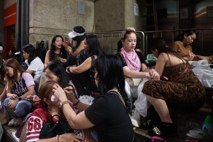 Groups of women, some giving manicures and makeup to each other, sit closely together on steps in an urban area.