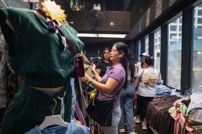 Women shop and browse through clothing at an indoor market stall, with tables and racks of clothes around them.