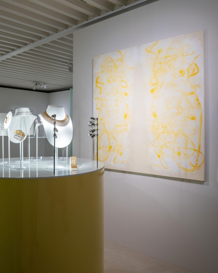 A jewellery display with gold necklaces and bracelets on stands, next to a large abstract yellow and white painting on the wall.