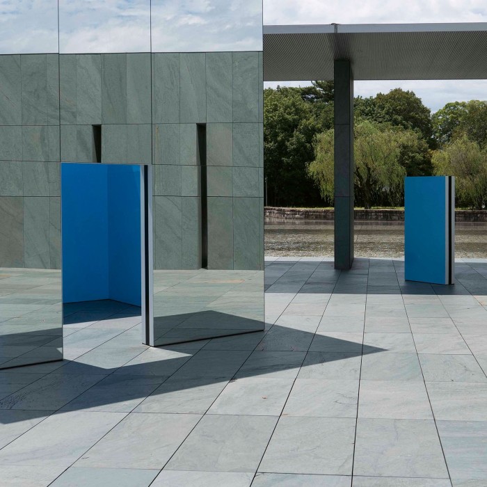 Outdoor installation featuring mirrored and blue geometric panels arranged within a minimalist stone courtyard surrounded by trees.