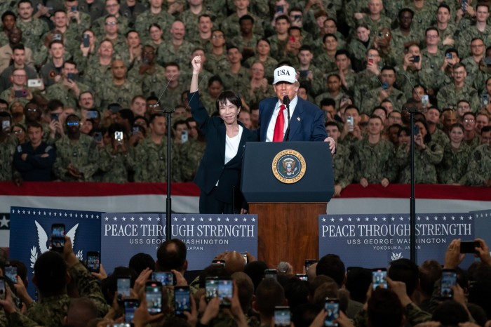 Japanese Prime Minister Sanae Takaichi raises her fist as U.S. President Donald Trump speaks at a podium aboard the USS George Washington, surrounded by uniformed personnel.