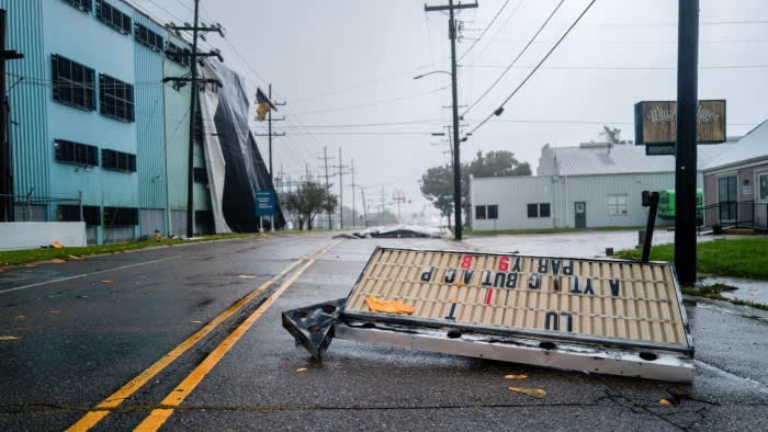 A storm-damaged street with a toppled sign lying across wet asphalt, flanked by leaning power lines and industrial buildings under grey skies