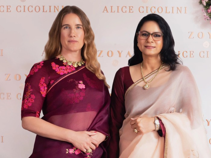 Two women in saris pose together at an event backdrop with the names Zoya and Alice Cicolini.