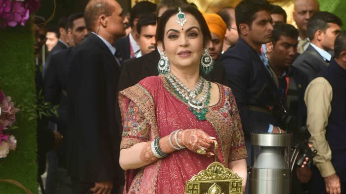 A woman dressed in a richly adorned pink saree with intricate embroidery poses gracefully. She wears heavy emerald and diamond jewellery and carries a golden clutch. Security personnel and attendees are visible in the background