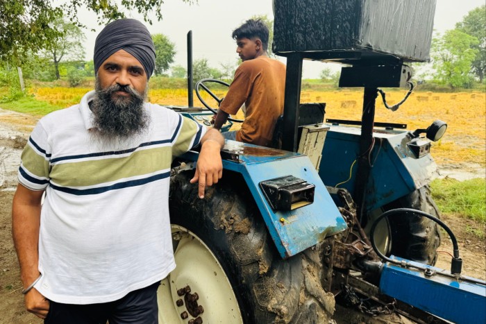 Daljinder Singh Haryaoo, wearing a grey turban, stands by a tractor in a field with another person seated on the tractor.