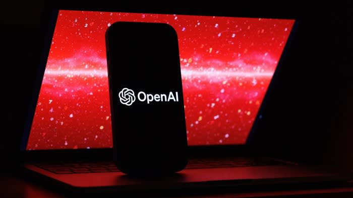 The OpenAI logo is displayed on a mobile phone screen in front of a laptop showing red digital graphics with numbers.