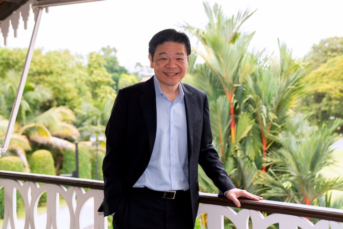 Lawrence Wong stands smiling on a balcony with lush greenery in the background, wearing a dark suit and light blue shirt.