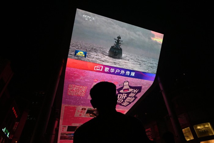 A man stands in silhouette watching a giant screen in Beijing that is showing footage of a Chinese military ship at sea.
