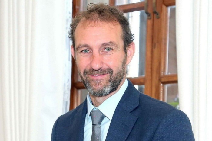 Matt Collins wearing a blue suit and smiling in front of a window with white curtains.