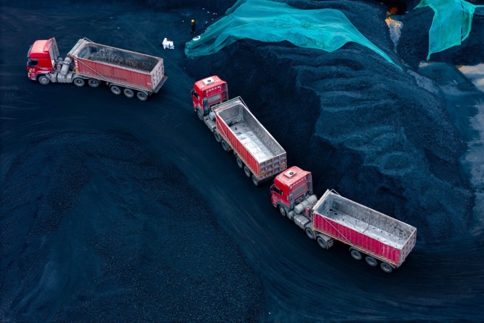 Three red trucks wait in line to be loaded with coal at a coal storage facility surrounded by large coal piles.