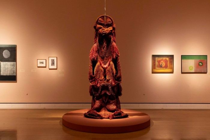 A large, textured fibre sculpture is displayed on a round platform, surrounded by paintings on a gallery wall