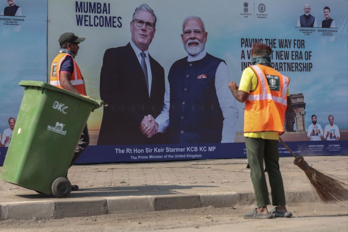 Posters of Keir Starmer and Narendra Modi shaking hands are displayed on a roadside in Mumbai as two workers clean the street in front.