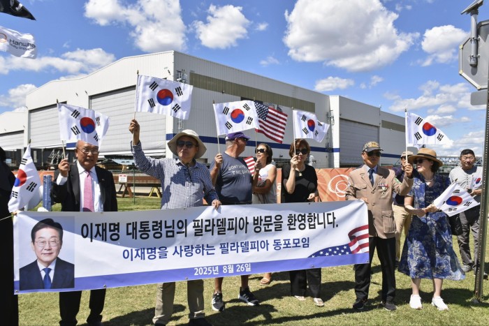 A group of people hold South Korean and US flags and a banner with a photo of South Korean President Lee Jae Myung outside Philly Shipyard.