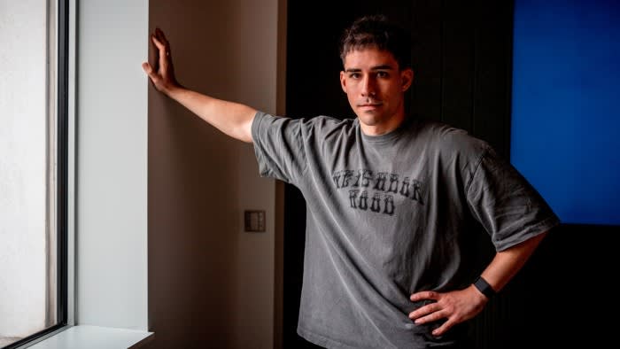 Alex Tkachenko stands indoors with one hand on his hip and the other resting against a window frame.