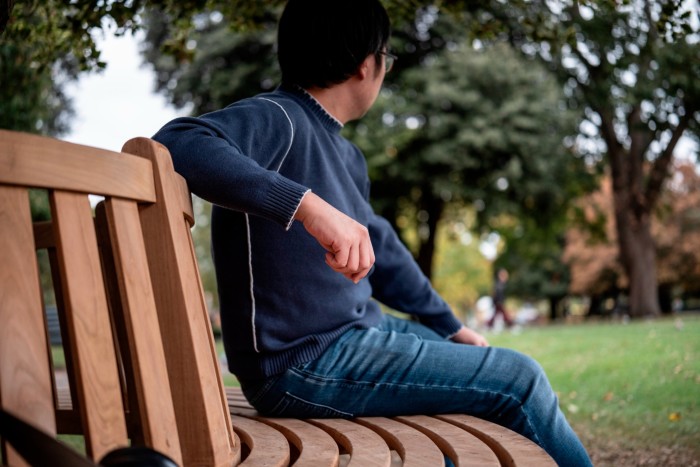Matthew sits on a park bench, turned away from the camera, wearing a blue sweater and jeans.