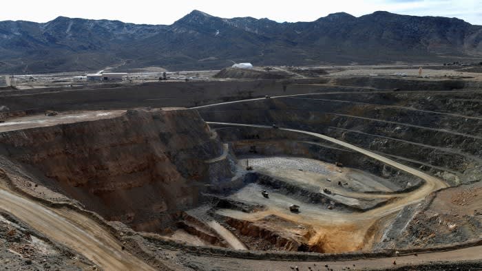 An open-pit rare earth mine with terraced walls and mining vehicles at the bottom, surrounded by mountains.
