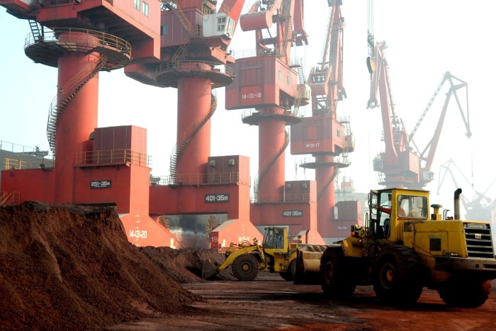 Bulldozers move piles of soil containing rare earth elements near large red cranes at an industrial port.