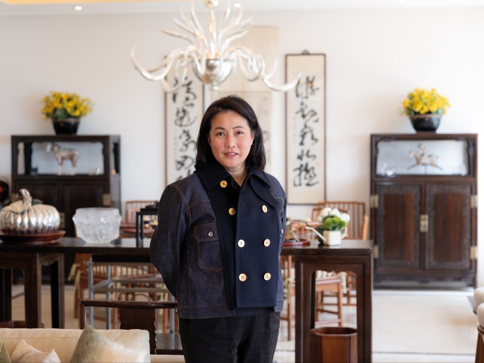 Patti Wong stands in an elegant living room with Chinese furniture, calligraphy scrolls and yellow flowers arranged symmetrically behind her.