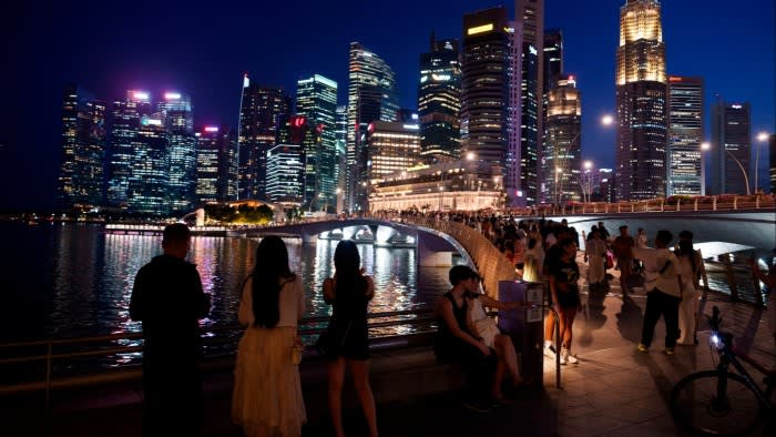 People gather along a waterfront promenade at night with Singapore’s illuminated skyline in the background
