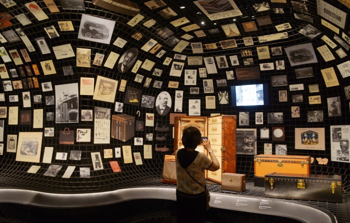 A woman photographs vintage Louis Vuitton trunks displayed in front of a curved wall covered with historical photos and documents.