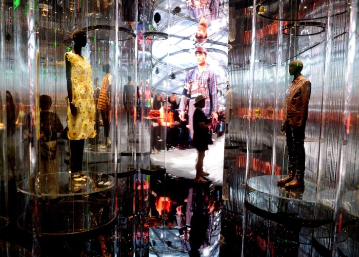 Mannequins dressed in designer clothing are displayed in transparent cylinders, with visitors and a large fashion image reflected throughout the multisensory Louis Vuitton flagship.