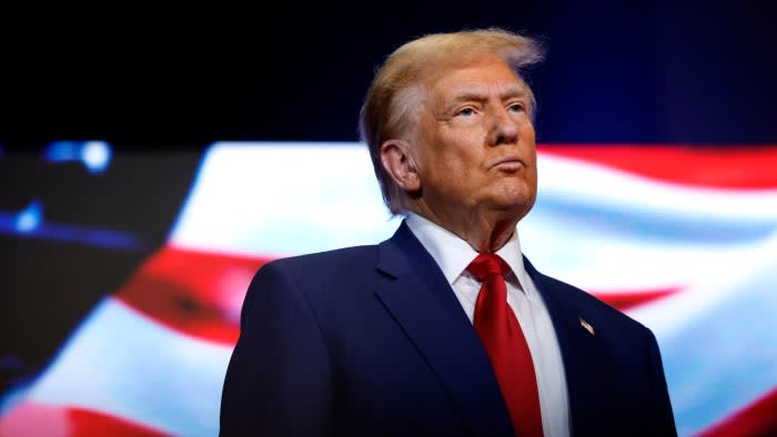 Donald Trump looks forward during a roundtable with faith leaders, with a blurred American flag in the background.
